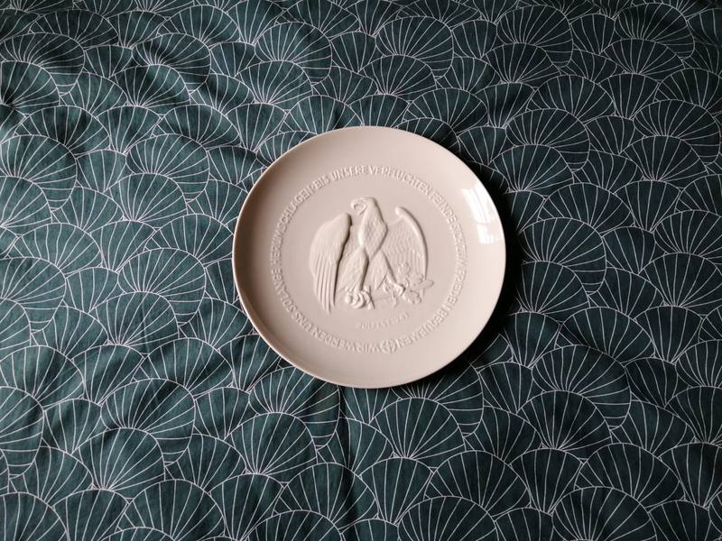 Une assiette blanche en porcelaine, ornée d'un aigle en relief, posée sur un tissu vert.