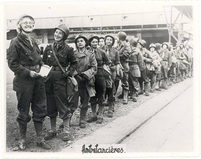 Une longue file de femmes casquées sur un quai ; en tête, Florence Conrad rit aux éclats. Sur le tirage, écrit à la main : Ambulancières.